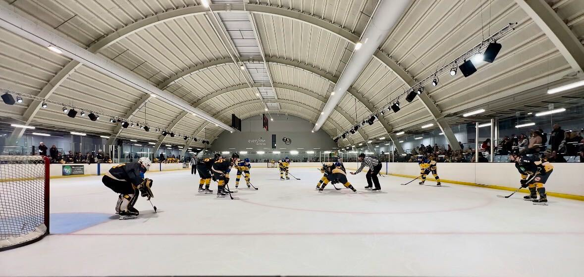 A wide indoor hockey rink with players lined up for a faceoff under arched arena lights